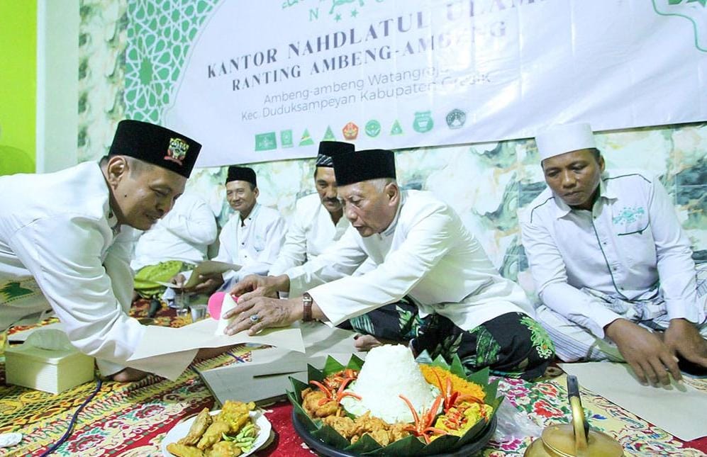 Rais Syuriyah MWCNU Duduksampeyan KH. Ach. Khusairi menyerahkan potongan tumpeng kepada Ketua PRNU Dusun Ambeng-ambeng Ustadz Umar Tajuddin, di acara peresmian Kantor Ranting NU Dusun Ambeng-ambeng, Ahad (5/1/2025) malam. Foto: dok PRNU Dusun Ambeng-ambeng/NUGres